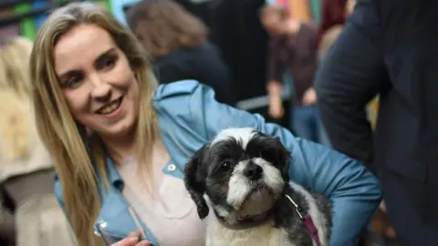 PA Media A young woman is pictured holding her black and white dog.
She has long blonde hair and is wearing a pale blue jacket over a light coloured top.
She is smiling.