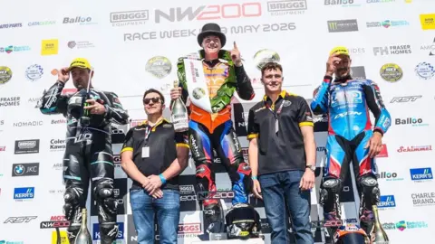 PACEMAKER Three male superbike riders standing on podiums in first, second and third place, in front of a big sponsors board showing it is the North West 200 race week. Two male support staff are also standing alongside. The winner Storm Stacey is standing on the number one podium, smiling and wearing a top hat and holding his index finger (pointer finger) up to show that he is number one. He's also holding a gold bottle of champagne and has a winners trophy, saying N.W 200 on it alongside his helmet, by his feet. It is a bright sunny day.