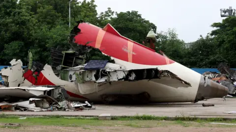 Part of the wreckage of the Air India Boeing 787-8 Dreamliner plane sits on the open ground. There is debris around as well as wiring.