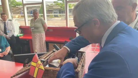 Laura Coffey/BBC Nigel Farage with short dark hair wearing glasses and a blue jacket accepts a brown wicker hamper containing a red and yellow Northamptonshire flag and locally-made wine and jam. A man has his hard in the basket, showing Farage around the contents. There are people standing in front of a red bench in the background, with a window behind them.