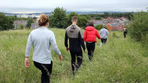 Pacemaker Five people dressed casually are seen from behind walking in single file through a grassy area close to houses
