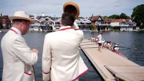 PA Media Two men in white suits with their back to the camera, looking out over a river where a team of rowers are sitting in a boat moored next to a pontoon. Across the river is a row of boat houses.
