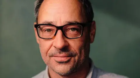A publicity shot of Paul Maltby. He has grey hair and stubble on his face and is wearing black-rimmed glasses.