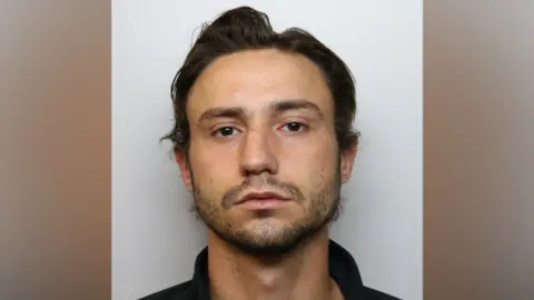 A man with dark hair and a beard appears in a police mugshot. He is wearing a black polo shirt.