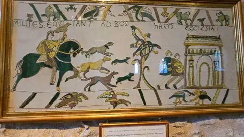 Fiona McCarthy A tapestry depicting a horseman, with an eagle resting on his arm. There are dogs and two men in front of him. 