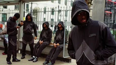 Dode Clothing Dimeji is in the foreground on the right in front of a bus stop where his friends are sitting in the background, all wearing black tracksuits. Mr Ode looks to the right of the frame, from under the hooded part of the top. His friends, three of them have the hoods pulled up, two of them sitting on the bench at the stop. The other two stand and both look at their phones.