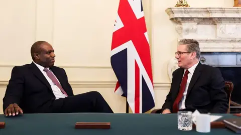 Prime Minister Keir Starmer sits, talking with Foreign Secretary David Lammy inside No10, in front of a Union Jack flag