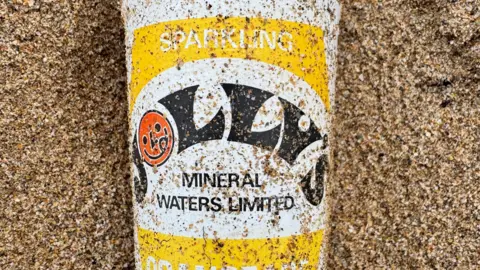 A white drinks can with black and red logo and yellow branding. It is placed on the sand.