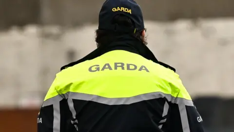 A garda officer photographed from behind.  They are wearing a navy cap and a navy jacket with fluorescent shoulders and reflective silver stripes