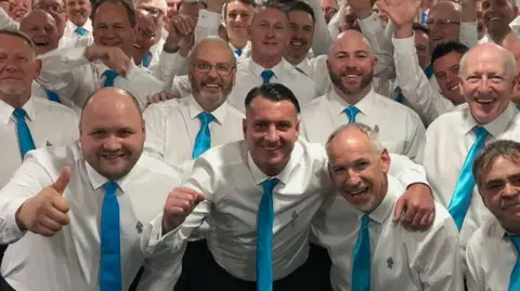 Peterborough Sings! A number of men are pictured together smiling and wearing white shirts and blue ties. They are arm-in-arm.