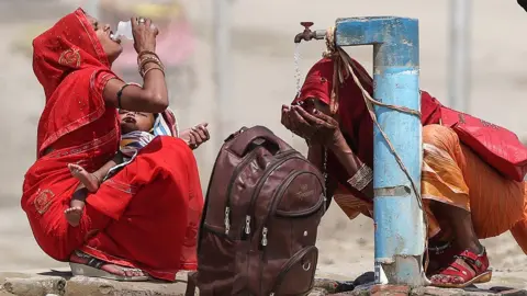 Two women drink water and splash their face from a public tap on a street in Prayagraj, India, on 10 June. They both wear red, with one holding a baby and a backpack on the ground next to them.