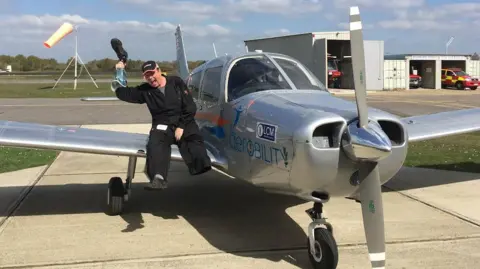 supplied A man in black overalls and a baseball cap sits on the wing of small plane. He is smiling and holding up a prostetic leg