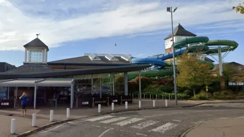 The entrance to Leisure World. It is a modern looking building with a mostly glass facade. However, off to the right is a tower from which a green and a blue flume come out of and lead down towards the back of the building.