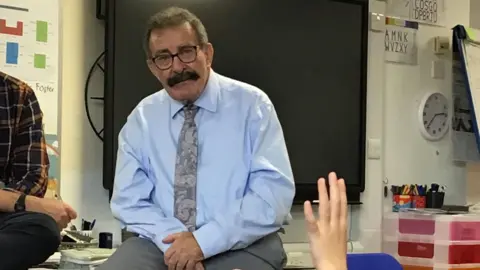 Lord Winston with dark hair and beard, wearing a light blue shirt and grey tie, sitting on a desk in a classroom. There is a large TV screen behind him. A hand is visible, raised by a child who is otherwise out of shot.