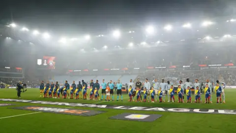 PA Media Maccabi Tel Aviv players in green tracksuits and Aston Villa in light blue tracksuits line up on the pitch with mascots. Behind them is a stand that is full on one side with home fans but empty on the left side where the away fans could have been before the ban. 