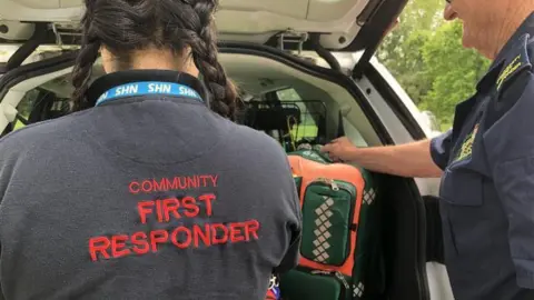 A woman and a man stand facing a car boot and medical equipment. The words Community First Responder are written on the back of the woman's jumper in red letters.