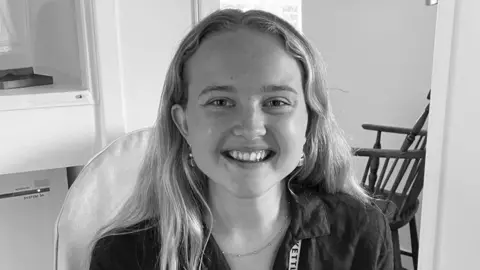 Supplied Black and white images of Letty Pilgrim smiling at the camera. She has long light hair, earrings, necklace and wears and shirt.
