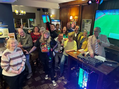 Geoff King A crowd of people stand in a pub with Geoff and Val King. He is speaking into a microphone behind a desk with audio mixing equipment on