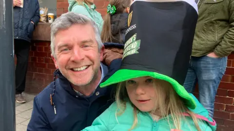 A man with short grey hair wearing a blue raincoat smiles at the camera. He is holding his daughter, a young girl with long blonde hair wearing a green coat and black top hat with a Guinness logo