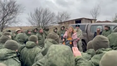 T.ME/KRSOLDATA Woman in pink coat playing guitar, surrounded by men in military uniform