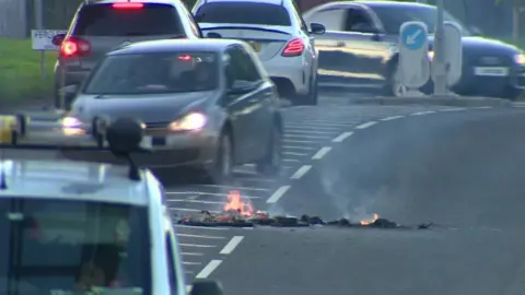 BBC Cars drive past burning debris on a road in Galliagh in Londonderry