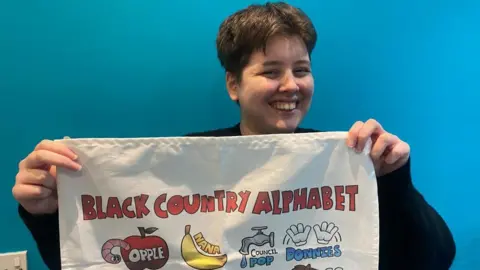 A lady with short brown hair, wearing a black jumper, is holding up a white tea towel to the camera. The tea towel reads "Black Country alphabet" and has drawings of an apple, banana, tap and pair of white gloves. The lady is smiling into the camera. 