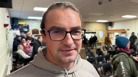 Andrew Turner/BBC Jason Lomasz has long dark hair tied back, is wearing black rimmed spectacles, and a grey hoodie. He is standing in the village hall at Hemsby, with other people and rows of chairs visible around him.