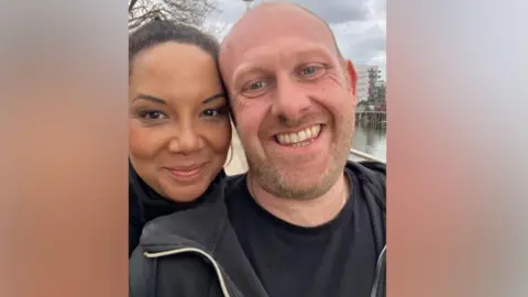 Supplied Caz (left) and Stock are smiling to the camera in a selfie. Caz has her hair tied back and is resting her head against Stock who is bald and has a ginger beard. They are both wearing black tops.