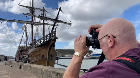 Robby West/BBC A man in a purple T-shirt who is holding a camera up and taking a picture of the wooden ship. The sky is blue and there are clouds above in the sky.