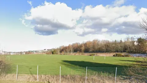 A large grassy playing field behind a tall wire fence at Inverness Royal Academy, with a blue sky and fluffy clouds.