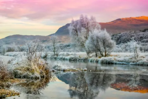 John Dewar Landscape image of a pale pink sky over the hills with frost-covered trees and grass below next to a river