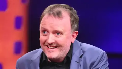 PA Media Comedian Chris McCausland, wearing a black shirt and powder blue jacket, smiles at someone or something off-camera.