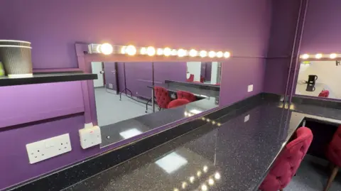 A view of a theatre dressing room with purple walls, upholstered red chairs, a marble-effect counter top, and a mirror lined with large light bulbs.