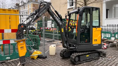 A yellow and black Volvo electric excavator on a road surrounded by barriers and metal-corrugated green and yellow portable cabins. 