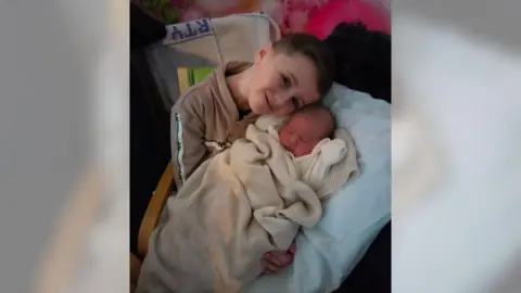 Medway NHS Foundation Trust Archie pictured holding his baby brother Alfie who was born on Christmas Day. Alfie is sleeping, swaddled in a dark cream blanket while Archie is wearing a cream hoodie. He leaning his head towards Alfie.