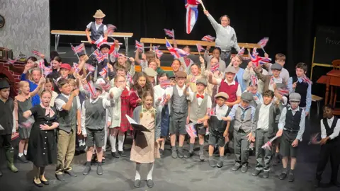 Schoolchildren are dressed in clothes from the 1940s as they sing on a stage with the light shining on them as they wave British and Jersey flags.