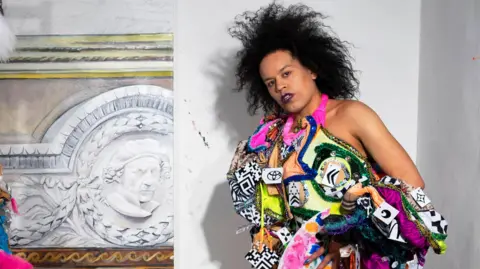 Quilla Constance Quilla, in a colourful outfit standing in a room but a piece of art. She is looking seriously at the camera. 