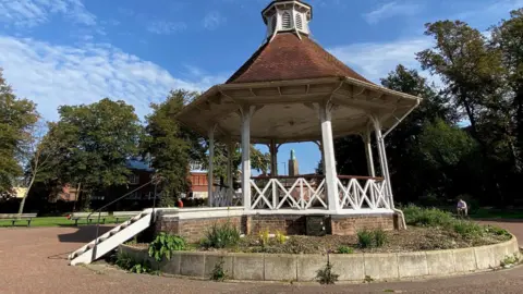 Norwich City Council A Bandstand on top of a stone base. The wooden structure has been painted white and has steps up to it. Around it are trees and benches. 