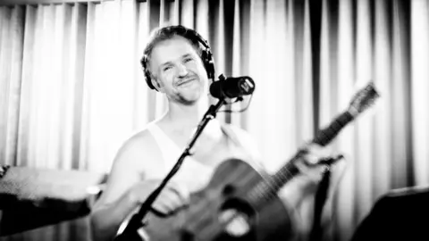 Luke Concannon A smiling Luke Concannon is standing in front of a microphone. He is wearing headphones and a white t-shirt and holding a guitar