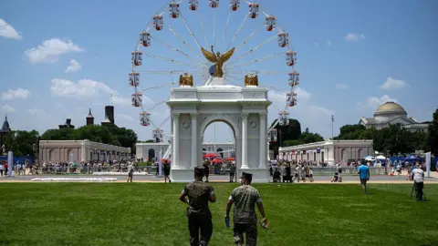 a view of two men walking through fair grounds in front of mock triumphal arch and ferris wheel