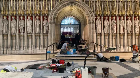 Workers are installing or repairing flooring inside a grand, ornate Gothic interior with detailed stone carvings and statues.
Tools, buckets, and equipment are scattered across the floor around a raised platform and steps.
The intricate backdrop features arched architecture and a stained-glass window, suggesting restoration within a historic building.