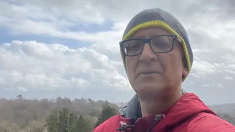 Dr Ravi Kumar taking a selfie whilst standing on a hill. He is wearing a green beanie and red jacket. Trees and bushes stretching into the horizon can be seen in the background.