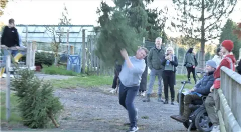 A man in a grey jumper and dark trousers throwing a Christmas tree over his shoulder.