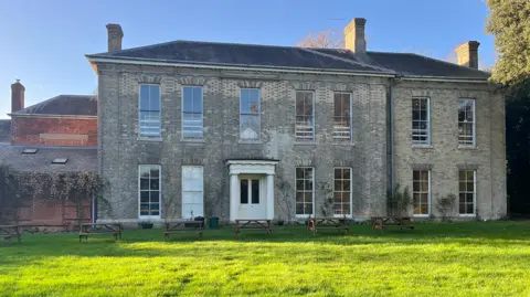 Clare Worden/BBC The front of Whitwell Hall, a two-storey manor house with large sash windows on both floors. The front door is framed by two pillars and white porch. There are a number of wooden picnic tables towards the front of the property and a red-brick extension to its side. 