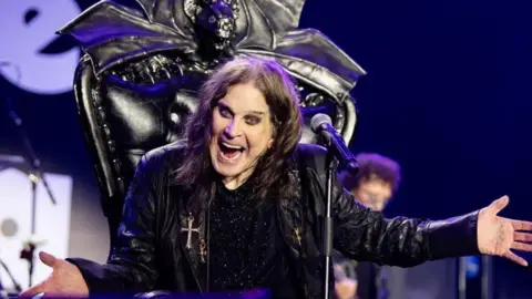 Ross Halfin Ozzy Osbourne on stage with his arms outstretched and smiling sat on a black leather throne with a microphone in front of him.
