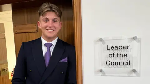George Finch leans on the door to the leader's office at Warwickshire County Council. He has short brown hair and smiles