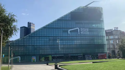 A general view of the National Football Museum, which is a glass building with a sloped roof. People can be seen walking past it. In the foreground is a park area with a path cutting through. 