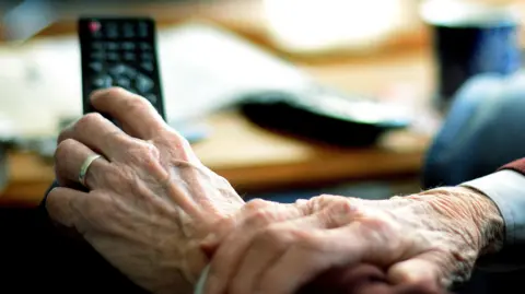 PA Media Close-up of the hands of an elderly person holding a television remote control. In the background there is a coffee table, upon which there is a coffee table, papers and other items.