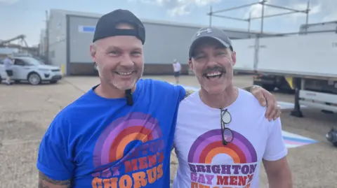 Josh McLaughlin/BBC Two men with their arms around each other. Both men are wearing t-shirts with a rainbow on them and the words: "Brighton Gay Men's Chorus".