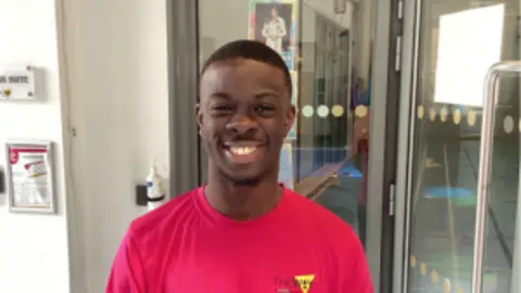 A man with short black hair and a goatee is wearing a dark pink tshirt and smiling widely at the camera, he appears to be standing in front of glass double doors